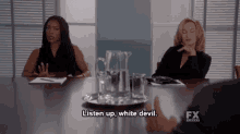 two women are sitting at a conference table and one of them says listen up white devil .