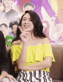a woman in a yellow off the shoulder top is smiling and giving a peace sign while sitting on a couch .