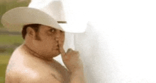 a shirtless man wearing a cowboy hat is holding his finger to his nose .