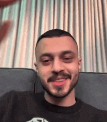 a man with a beard is sitting on a couch and smiling at the camera .