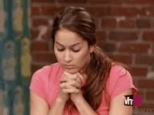 a woman in a pink shirt is sitting in front of a brick wall with her hands folded in prayer .