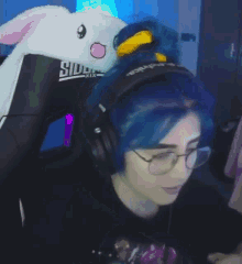 a woman with blue hair and glasses is wearing headphones and a stuffed bunny hat .