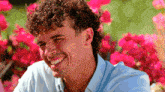 a man with curly hair is smiling in front of a bush of pink flowers