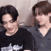 two young men are sitting next to each other on a couch . one of the men is wearing a shark shirt .