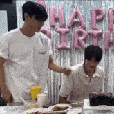 two men are sitting at a table with plates of food in front of balloons that say happy birthday .