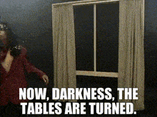 a woman standing in front of a window with the words now darkness the tables are turned .