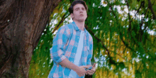a young man in a blue plaid shirt is standing under a tree holding a cell phone .
