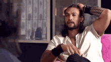 a man with long hair and a beard is sitting on a pink couch holding his hair .