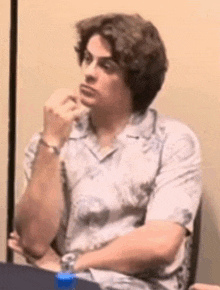 a man in a white shirt is sitting at a table with his hands on his chin .