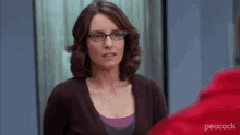 a woman wearing glasses and a brown sweater is standing in front of a red peacock logo .