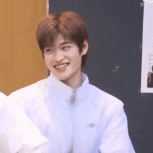 a young man in a white jacket is smiling while sitting in front of a wall .