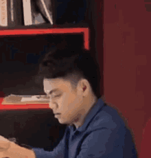 a man in a blue shirt is sitting at a table in front of a bookshelf and looking at his phone .
