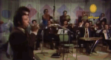 a man singing in front of an orchestra with arabic writing on the bottom