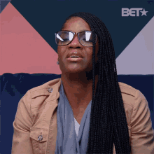 a woman wearing glasses and a tan jacket is sitting on a couch with a bet logo behind her