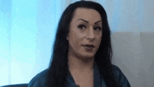 a woman with long black hair is sitting in a chair in front of a window and looking at the camera .