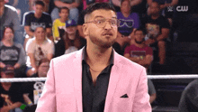 a man in a pink suit and glasses stands in a wrestling ring with a cw logo in the background