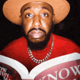 a man wearing a hat and a necklace is reading a book titled now