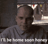 a bald man is sitting in front of a window with the words " i 'll be home soon honey " written below him .