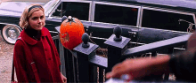 a woman in a red coat is standing next to a pumpkin on a porch .