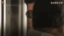 a man wearing sunglasses is standing in front of a screen that says sarkar