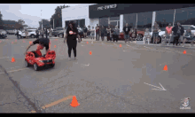 a man is driving a red toy car in a parking lot while a woman watches .