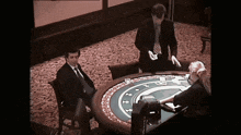 a man in a suit sits at a roulette table while another man stands behind it