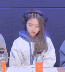 a girl wearing headphones sits at a table with a bottle of orange soda