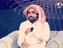 a man wearing a head scarf and a watch sits in a chair in front of a screen with arabic writing on it