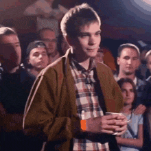 a young man in a plaid shirt is standing in a crowd of people holding a cup .
