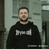 a man with a beard wearing a black hoodie with russian writing on it is standing in front of a building .
