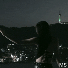 a woman is standing in front of a city at night with a green tower in the background and the letters ms on the bottom