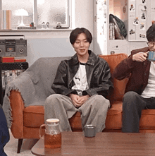 a man in a leather jacket is sitting on a couch next to another man drinking coffee .