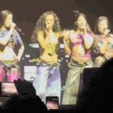 a group of women singing into microphones on a stage with a cell phone in the foreground