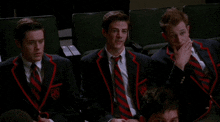three men in suits and ties are sitting in a auditorium