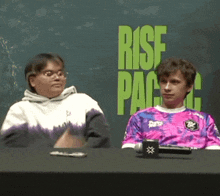 two people sit at a table in front of a wall that says rise pac