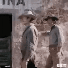 a couple of men standing next to each other in front of a brick wall .