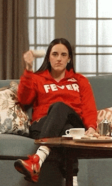 a woman in a red hoodie is sitting on a couch with a cup of coffee .