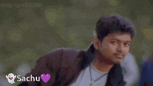 a man in a brown jacket is standing in front of a pink heart with the name sachu written on it .
