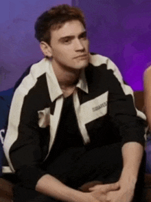 a young man wearing a black and white jacket is sitting on a couch with his hands folded .