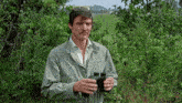 a man in a green jacket holds binoculars in front of a field