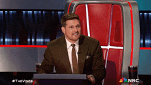 a man in a suit and tie is sitting in front of a podium with the nbc logo on it