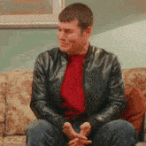a man wearing a black leather jacket and a red sweater sits on a couch