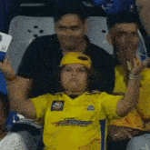 a young girl wearing a yellow shirt and a yellow hat is sitting in a stadium with her arms in the air .