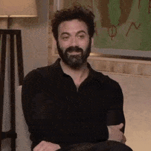 a man with a beard is sitting in a chair with his hands folded and smiling .