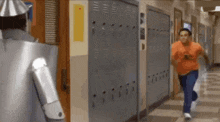 a man in an orange shirt is running down a hallway in front of a robot .