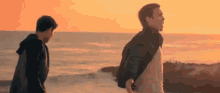 two men are walking on the beach at sunset with their backs to the camera .
