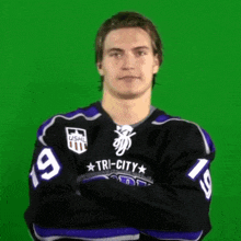 a man in a tri-city jersey stands with his arms crossed in front of a green screen