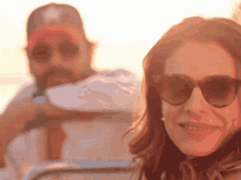 a woman wearing sunglasses smiles in front of a man wearing a baseball cap