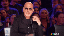 a bald man is speaking into a microphone in front of a crowd of people with a nbc logo in the background