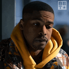 a close up of a man wearing a yellow hoodie with a be t logo behind him